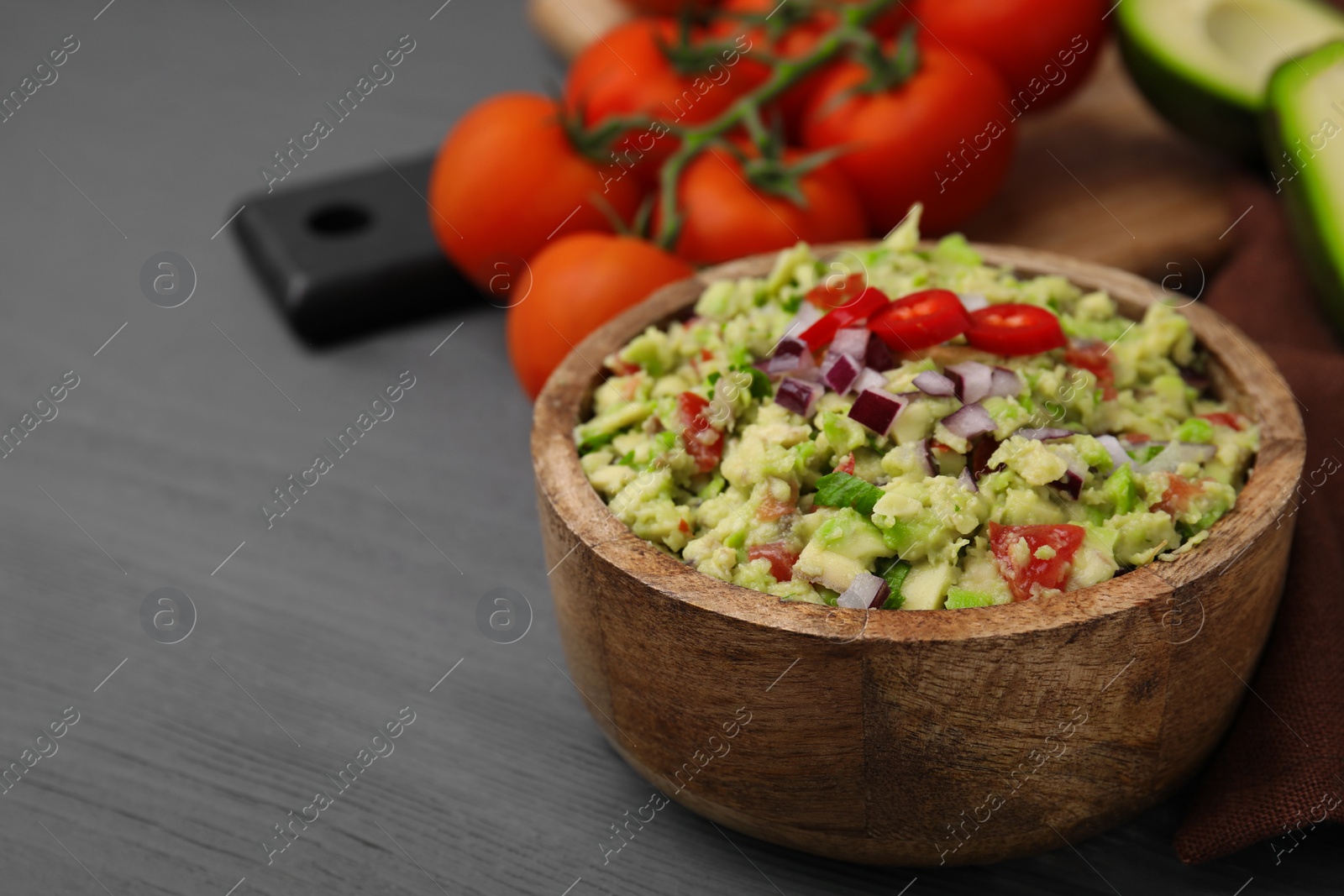 Photo of Bowl of delicious guacamole and ingredients on grey wooden table, closeup. Space for text