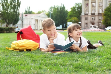 Photo of Children with school stationery reading book on green lawn outdoors