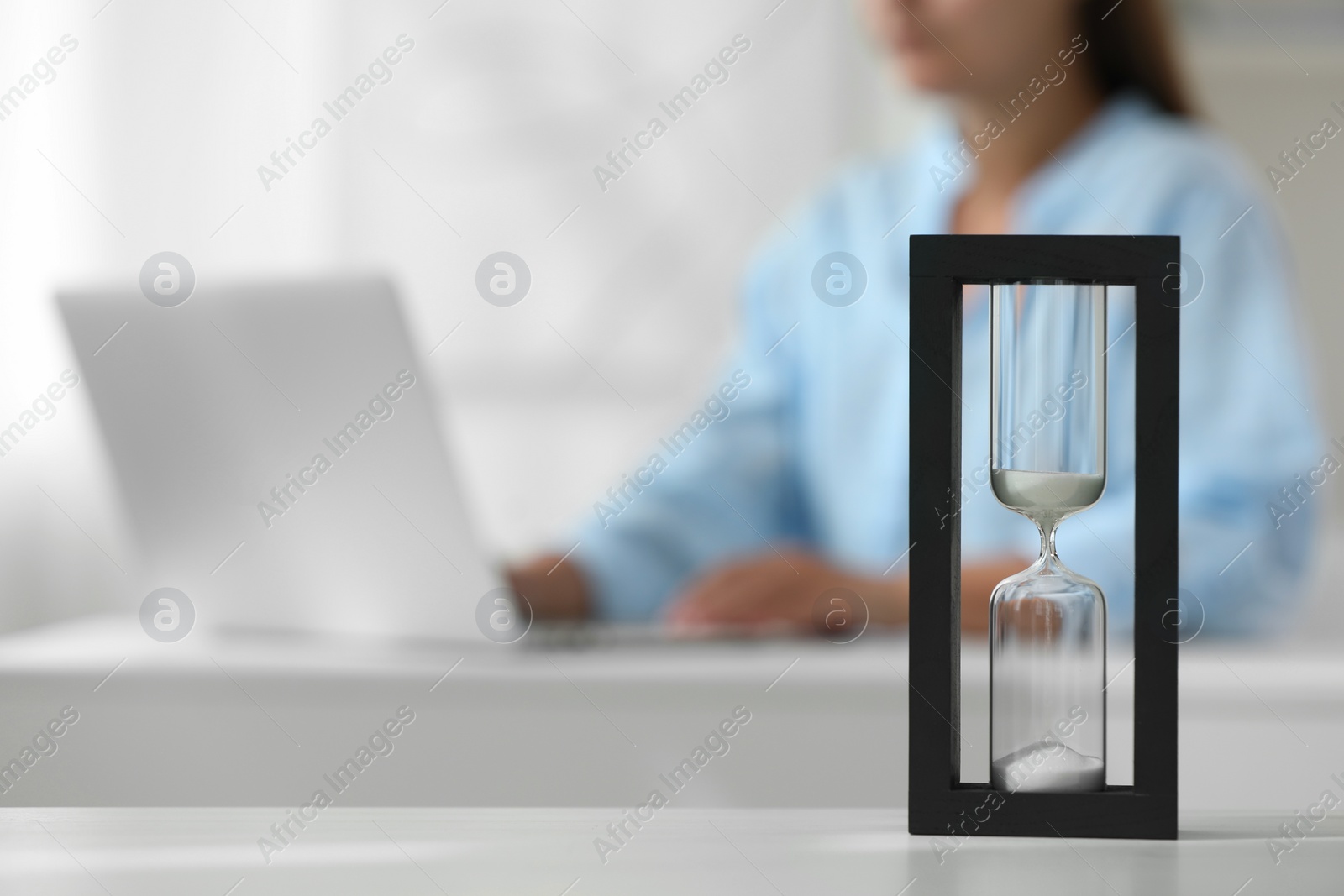 Photo of Hourglass with flowing sand on white table. Woman using laptop indoors, selective focus