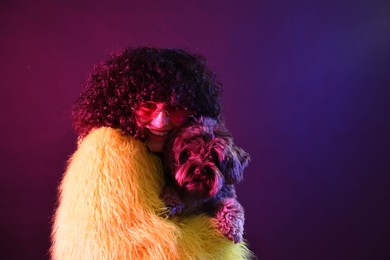 Photo of Beautiful young woman in yellow fur coat and sunglasses with cute dog on color background in neon lights. Space for text