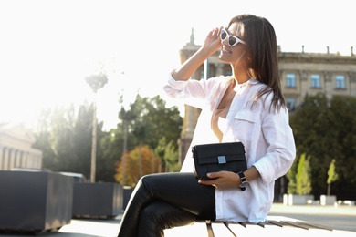 Young woman in sunglasses with stylish black bag on bench outdoors