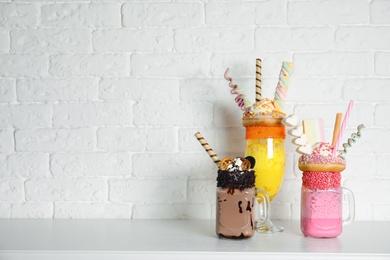 Tasty milk shakes with sweets in glassware on table near brick wall. Space for text