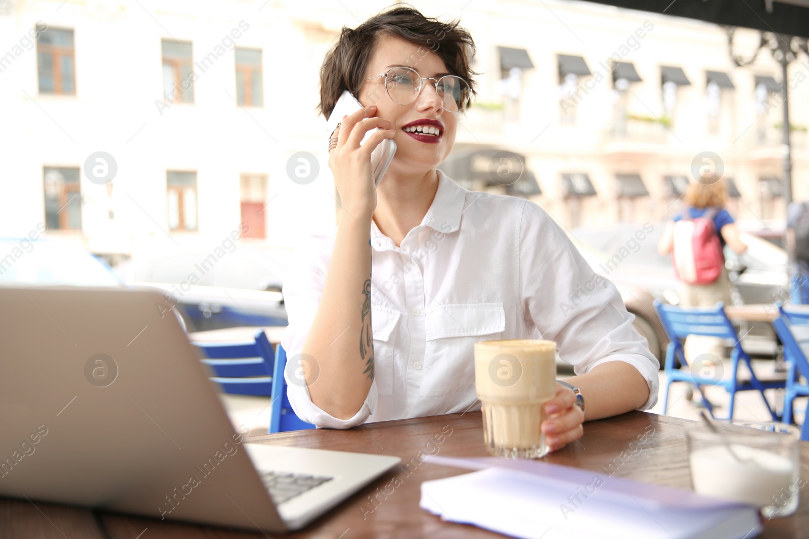 Photo of Young woman talking on phone while working with laptop at desk in cafe