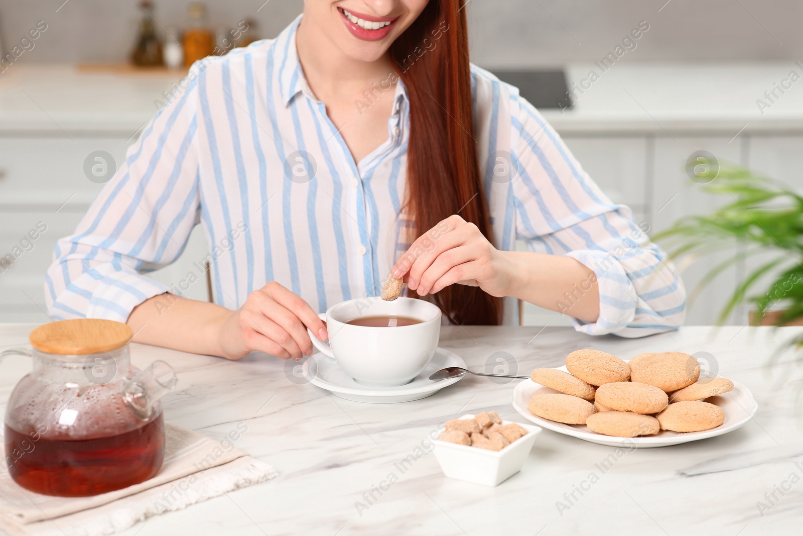 Photo of Woman with red dyed hair holding cookie above cup of tea at white marble table in kitchen, closeup