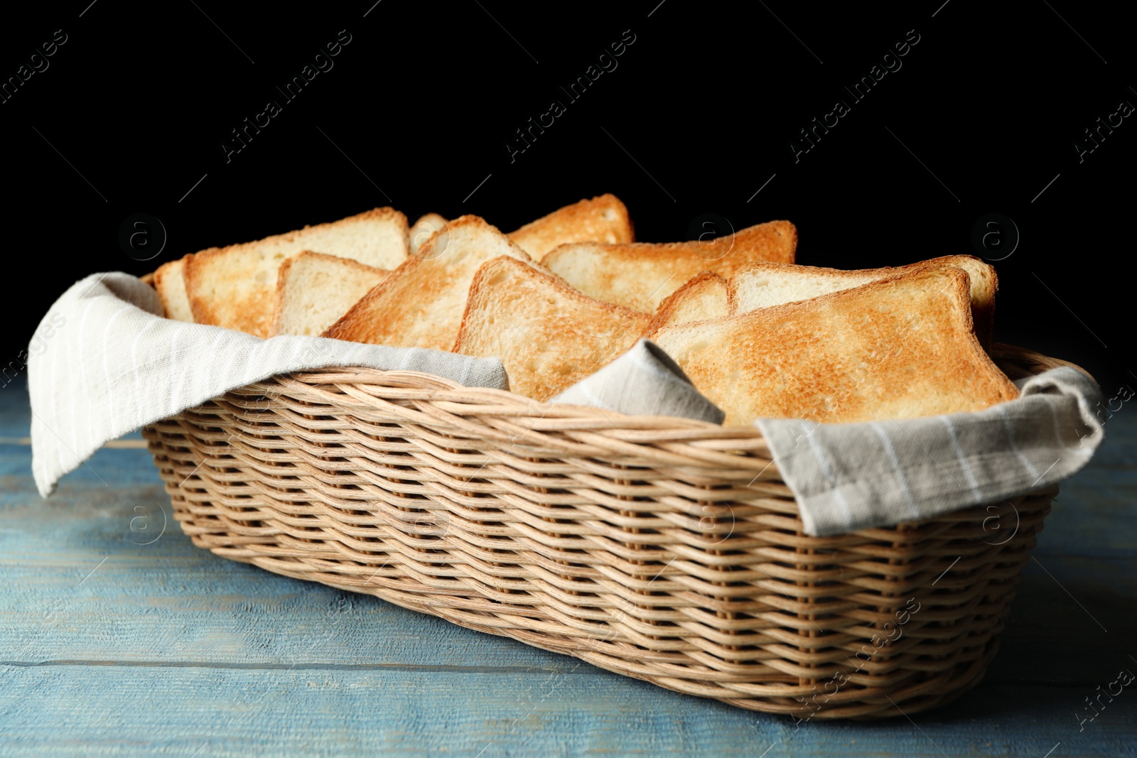 Photo of Slices of toasted bread in basket on light blue wooden table against black background