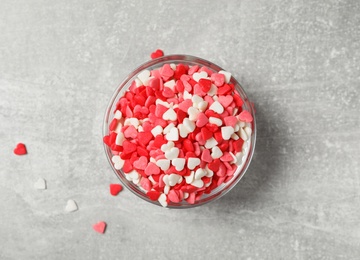 Bright heart shaped sprinkles in glass bowl on grey table, flat lay