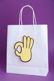 Paper bag and cutout of okay hand gesture on purple background