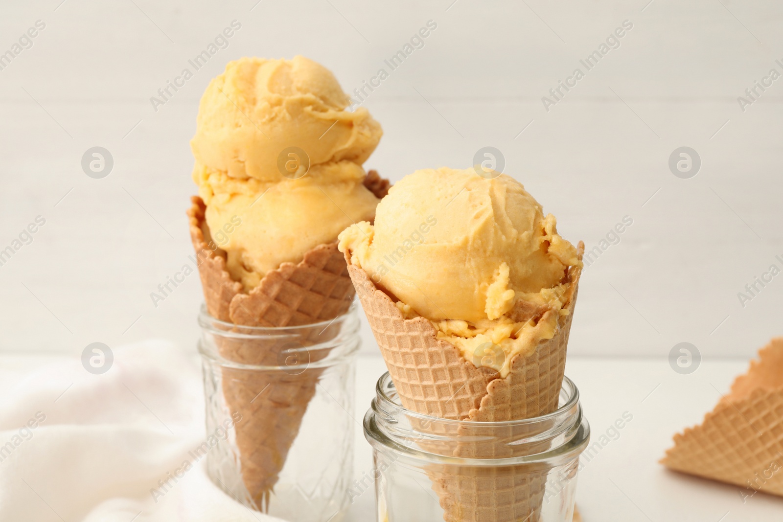 Photo of Delicious yellow ice cream in wafer cones and glass jars on table, closeup