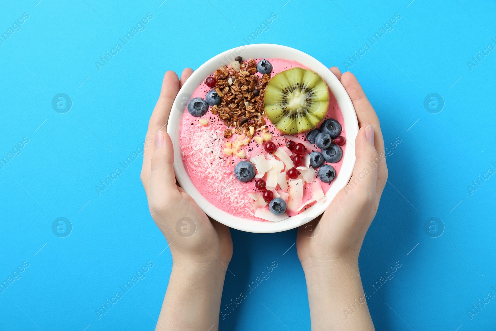 Photo of Woman holding tasty smoothie bowl with fresh kiwi fruit, berries and granola at light blue table, top view