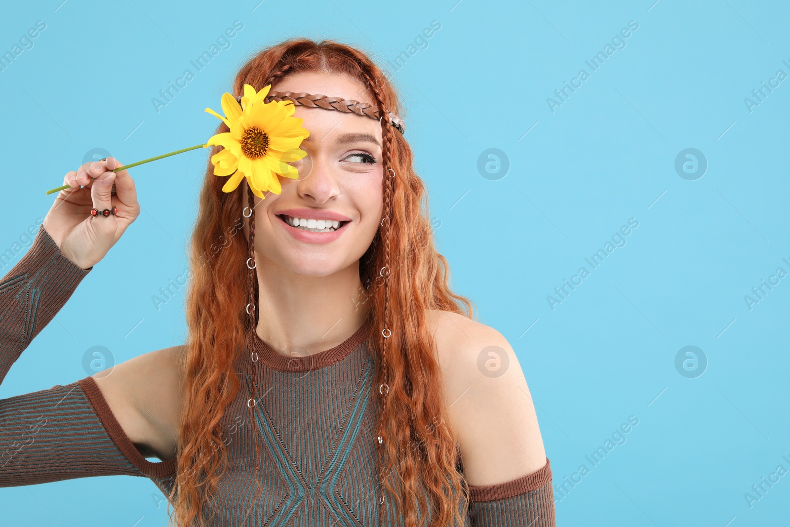 Photo of Beautiful young hippie woman covering eye with sunflower on light blue background, space for text