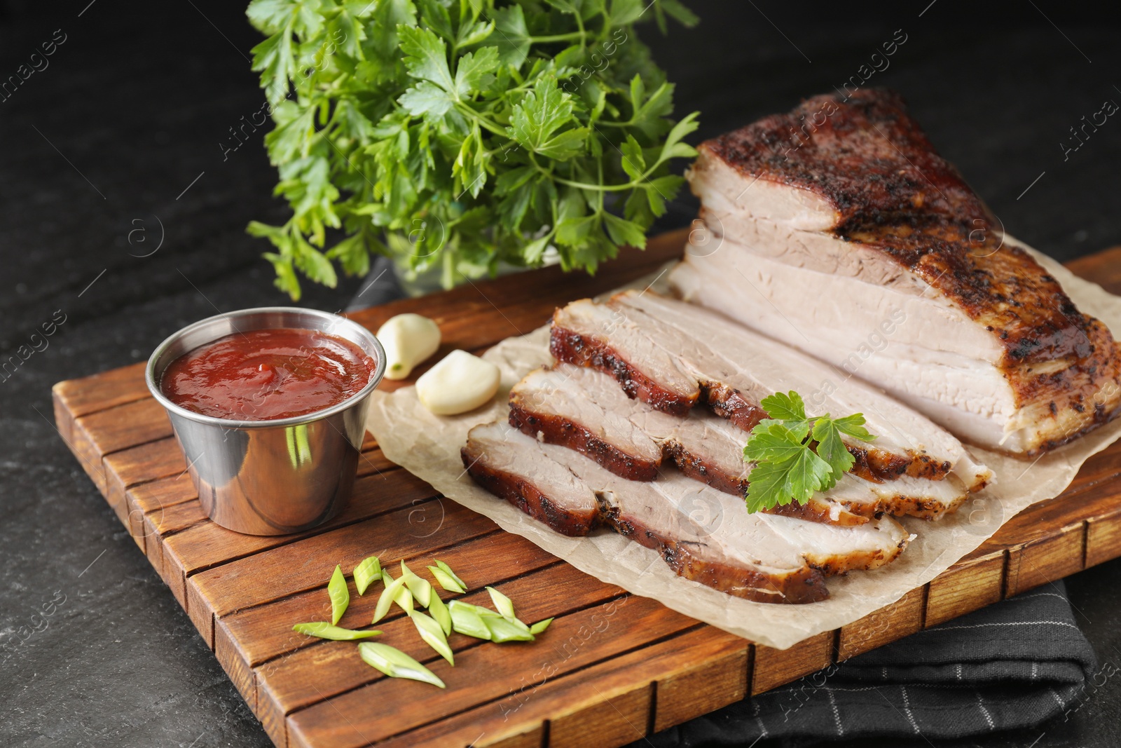 Photo of Pieces of baked pork belly served with sauce and parsley on black textured table, closeup