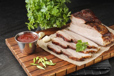 Photo of Pieces of baked pork belly served with sauce and parsley on black textured table, closeup