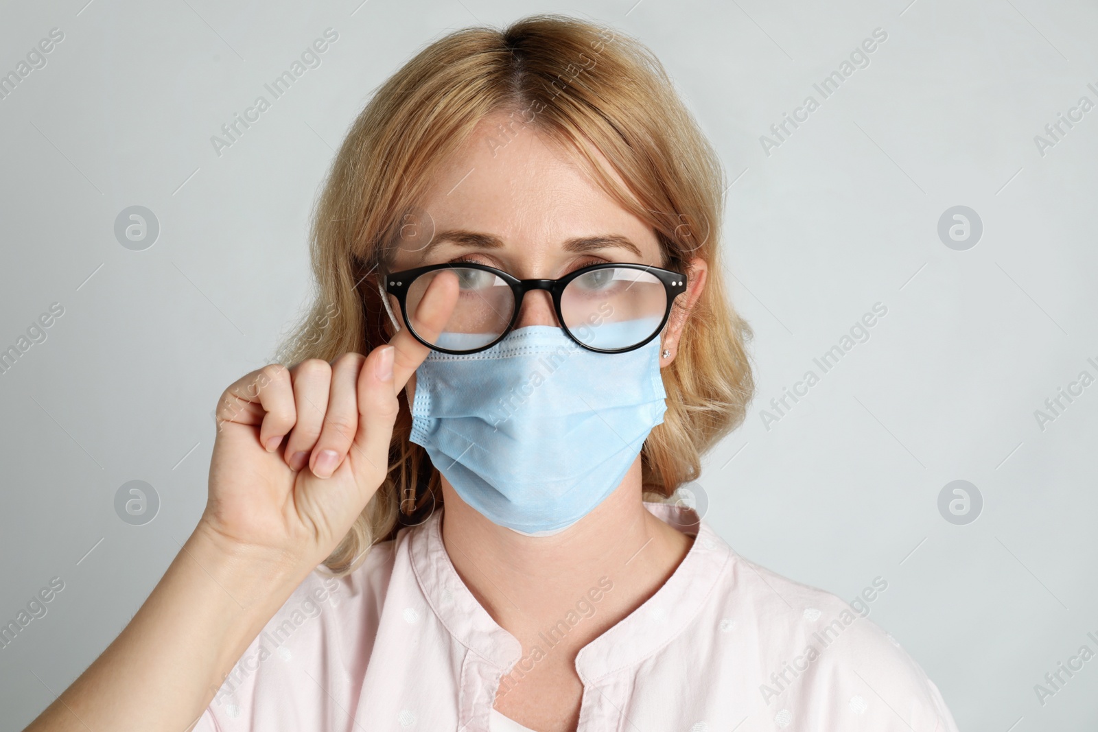 Photo of Woman wiping foggy glasses caused by wearing medical mask on light background