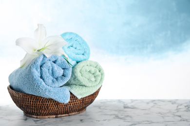Photo of Wicker basket with rolled bath towels and beautiful flower on white marble table. Space for text