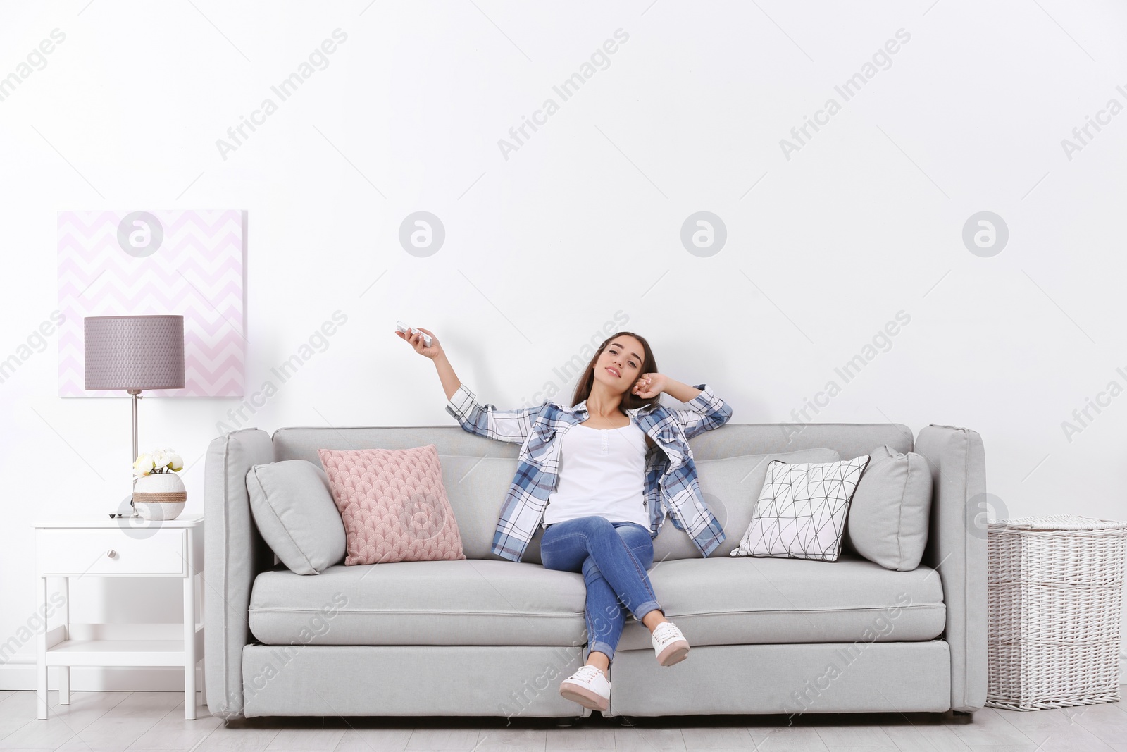 Photo of Young woman switching on air conditioner while sitting on sofa near white wall