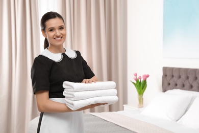 Young maid holding stack of fresh towels in hotel room. Space for text