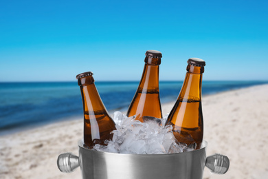 Image of Bottles of beer with ice cubes in metal bucket against blurred sea and sandy beach