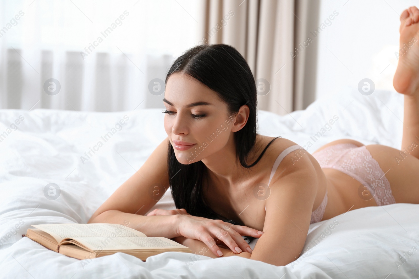 Photo of Beautiful young woman in pink underwear reading book on bed at home