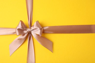 Beige satin ribbon with bow on yellow background, top view