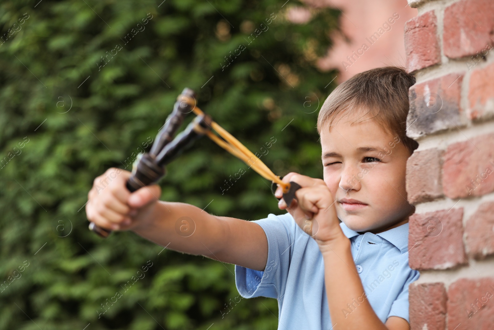 Photo of Little boy playing with slingshot near brick wall outdoors