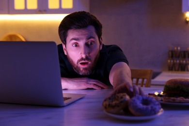 Photo of Man taking donut while using laptop in kitchen at night. Bad habit