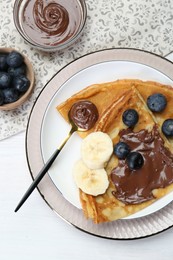 Photo of Tasty crepes with chocolate paste, blueberries and banana served on white wooden table, flat lay