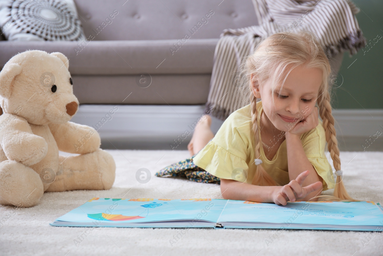 Photo of Cute little girl lying on cozy carpet with book and teddy bear at home