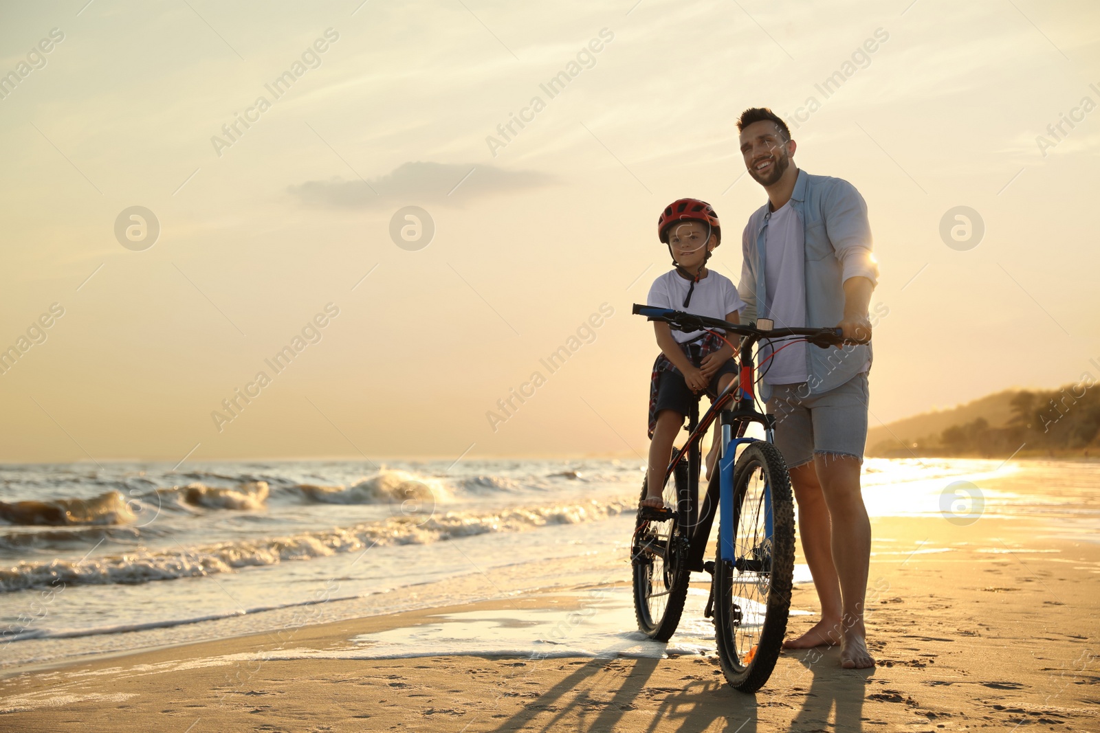 Photo of Happy father teaching son to ride bicycle on sandy beach near sea at sunset