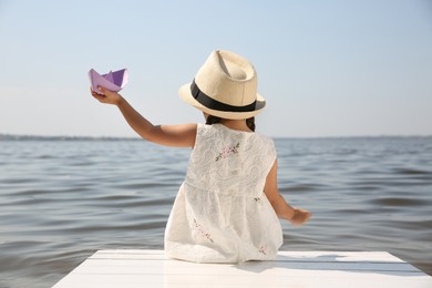 Photo of Little girl playing with paper boat on wooden pier near river, back view