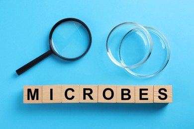 Word Microbes made with wooden cubes, magnifying glass and Petri dishes on light blue background, flat lay