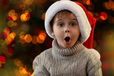 Photo of Emotional little boy in Santa Claus cap on blurred background. Christmas time