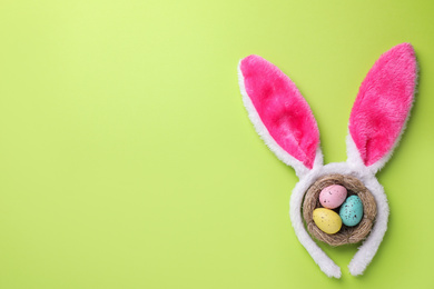 Headband with Easter bunny ears and dyed eggs in nest on green background, flat lay. Space for text