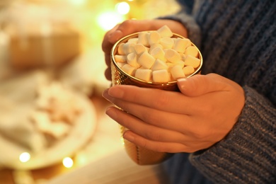 Woman holding cup of hot drink with marshmallows indoors, closeup. Magic Christmas atmosphere