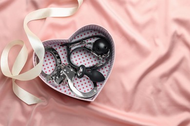Photo of Heart shaped gift box with different sex toys and ribbon on pink silk fabric, flat lay. Space for text