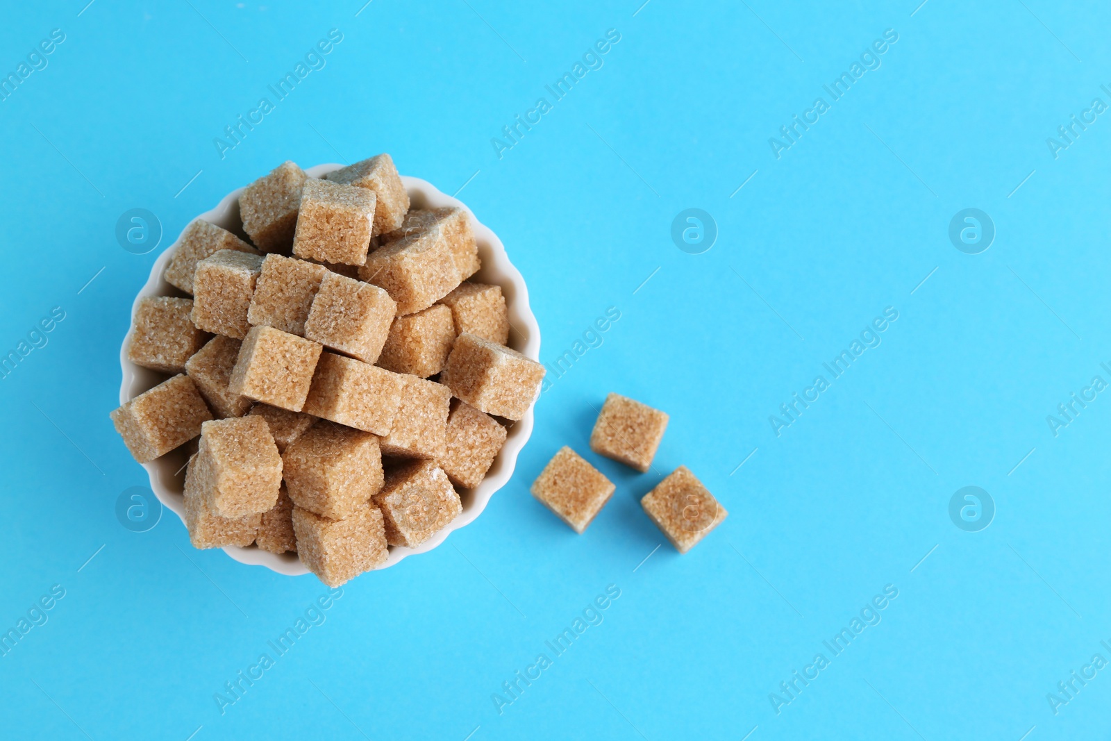 Photo of Brown sugar cubes in bowl on light blue background, top view. Space for text