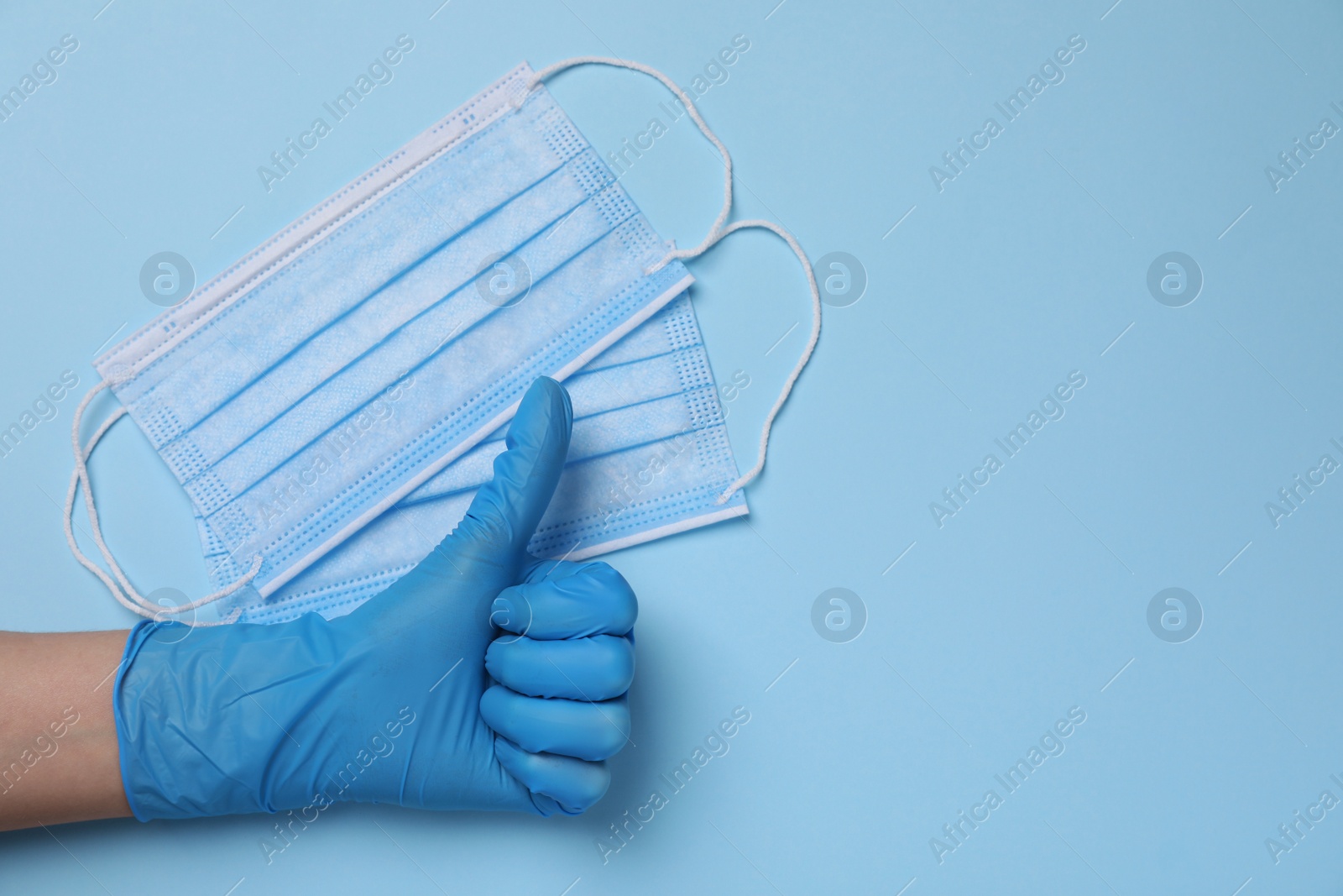Photo of Woman showing thumb up near protective masks on light blue background, top view with space for text. Safety equipment