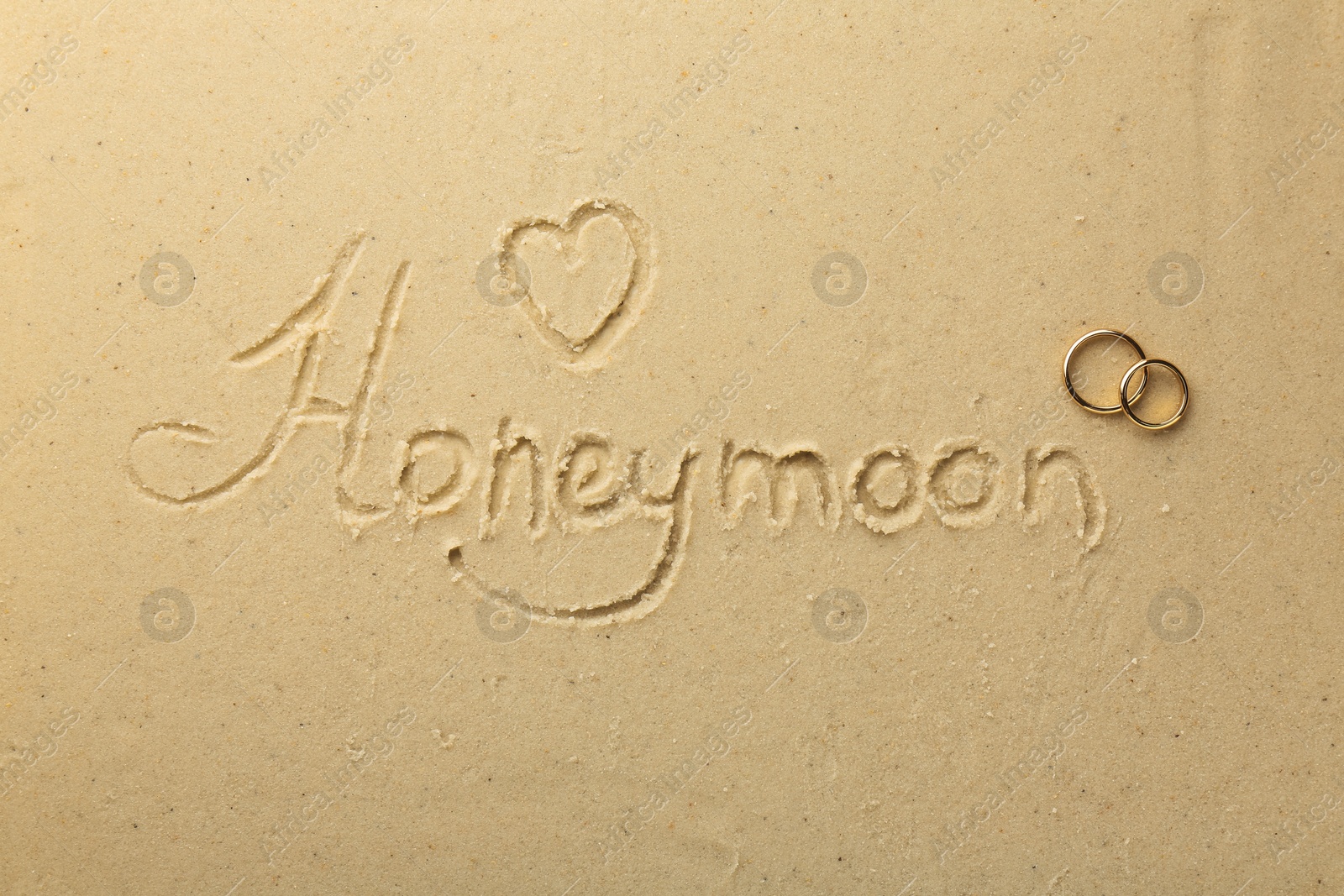 Photo of Word Honeymoon and heart written on sand and two golden rings, top view