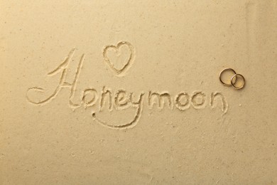 Word Honeymoon and heart written on sand and two golden rings, top view