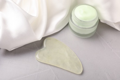 Jade gua sha tool , jar of cream and white fabric on grey table