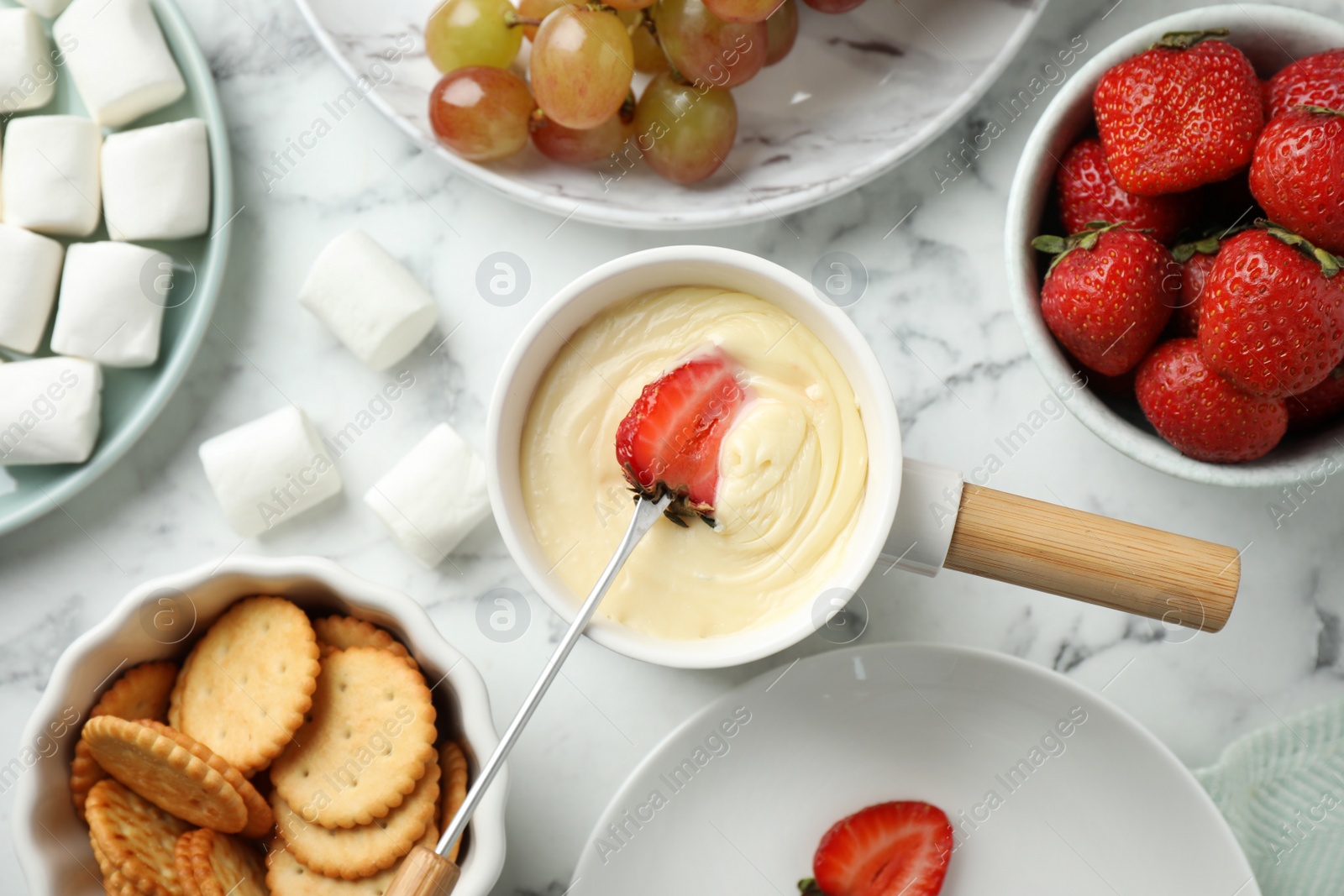 Photo of Dipping strawberry into fondue pot with white chocolate on marble table, top view