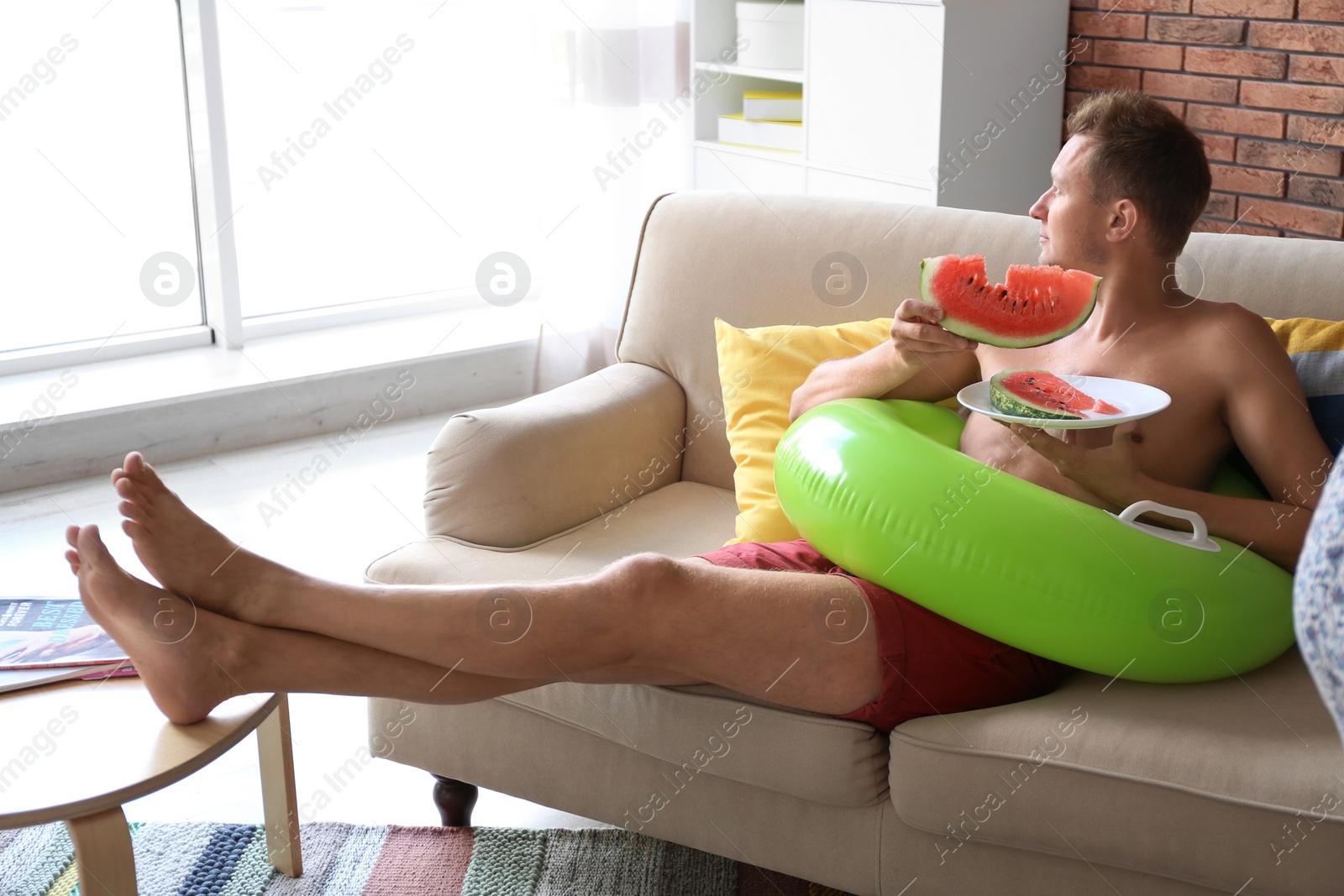 Photo of Shirtless man with inflatable ring eating watermelon on sofa at home