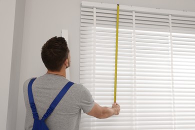 Photo of Worker in uniform using measuring tape while installing horizontal window blinds indoors