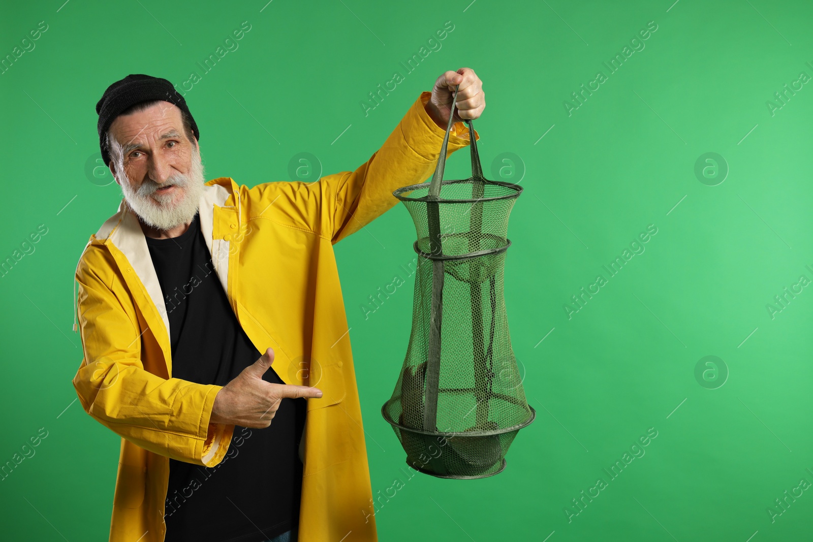 Photo of Fisherman holding fishing net with catch and pointing on green background, space for text