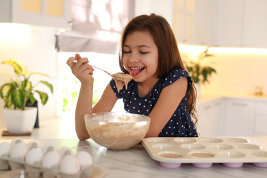 Photo of Cute little girl licking raw dough from spoon in kitchen. Cooking food