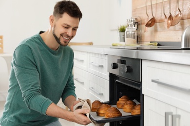 Photo of Handsome man taking out tray of baked buns from oven in kitchen