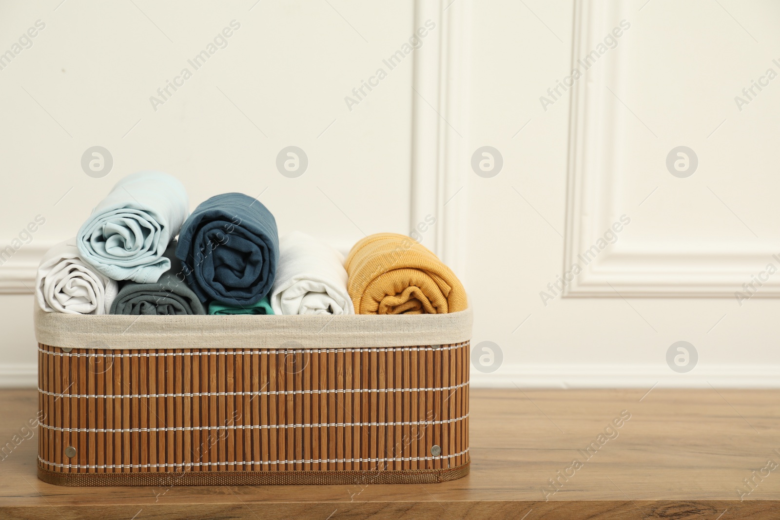 Photo of Basket with different rolled clothes on wooden table near white wall, space for text