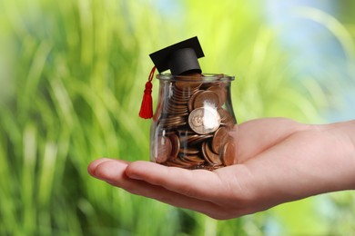 Photo of Man holding glass jar with coins and graduation cap against blurred background, closeup. Scholarship concept