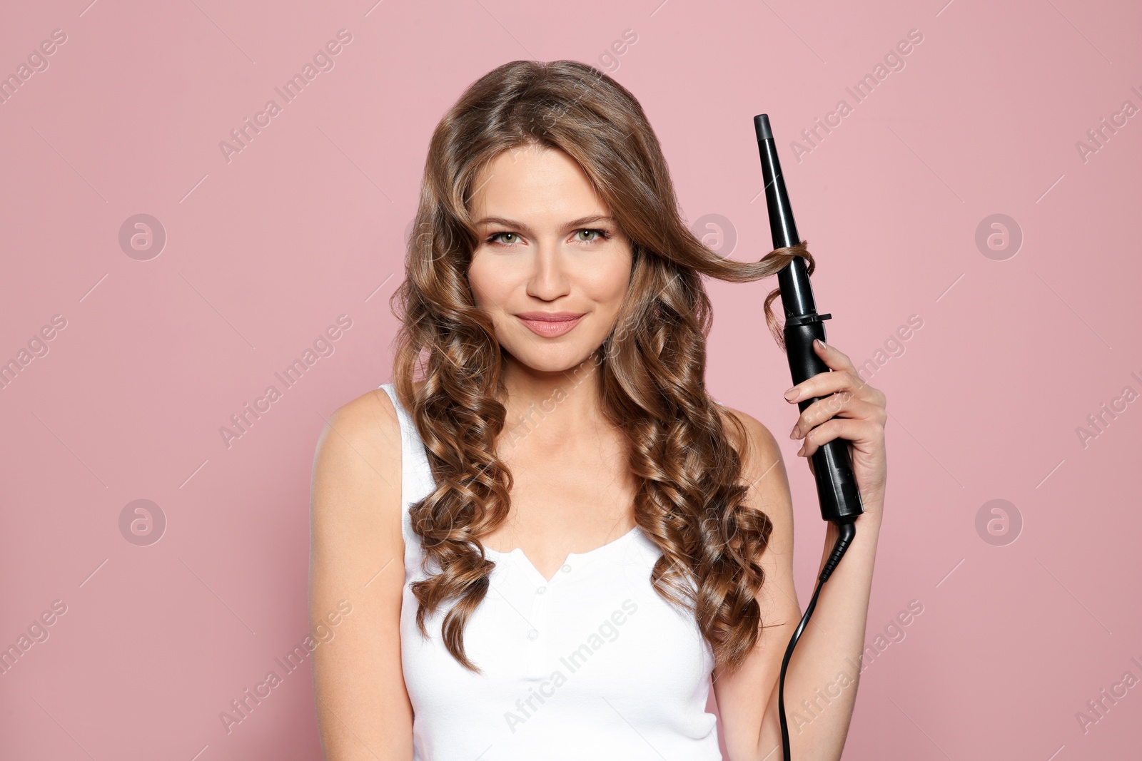 Photo of Portrait of young woman with shiny wavy hair using curling iron on color background