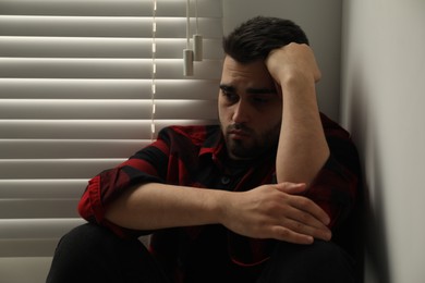 Sad young man near closed blinds at home