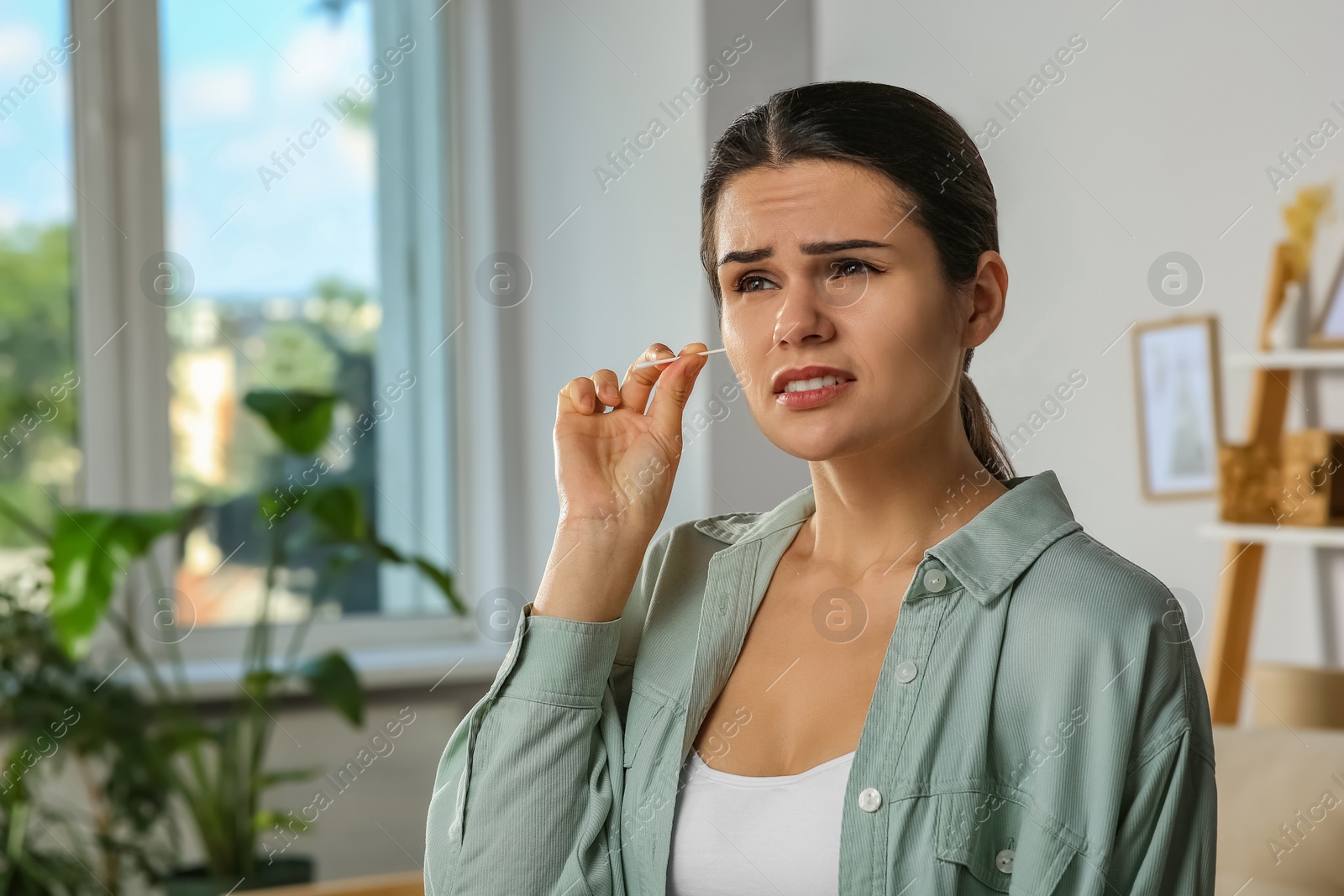 Photo of Young woman cleaning ear with cotton swab in room